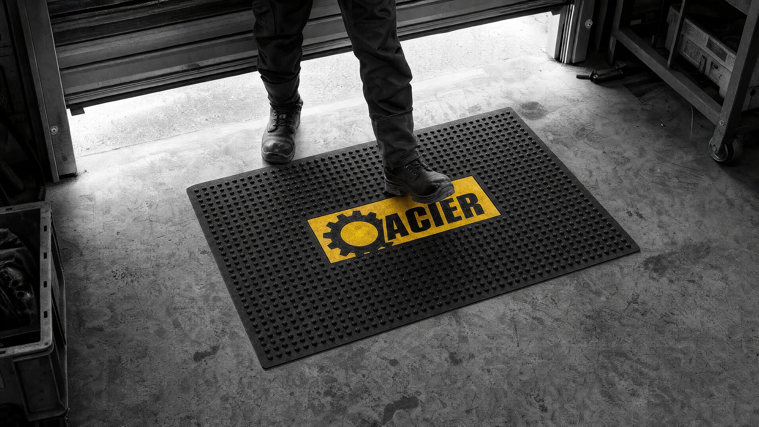 Tapis en caoutchouc noir à picots avec un logo jaune moulé, à l’entrée d’un atelier