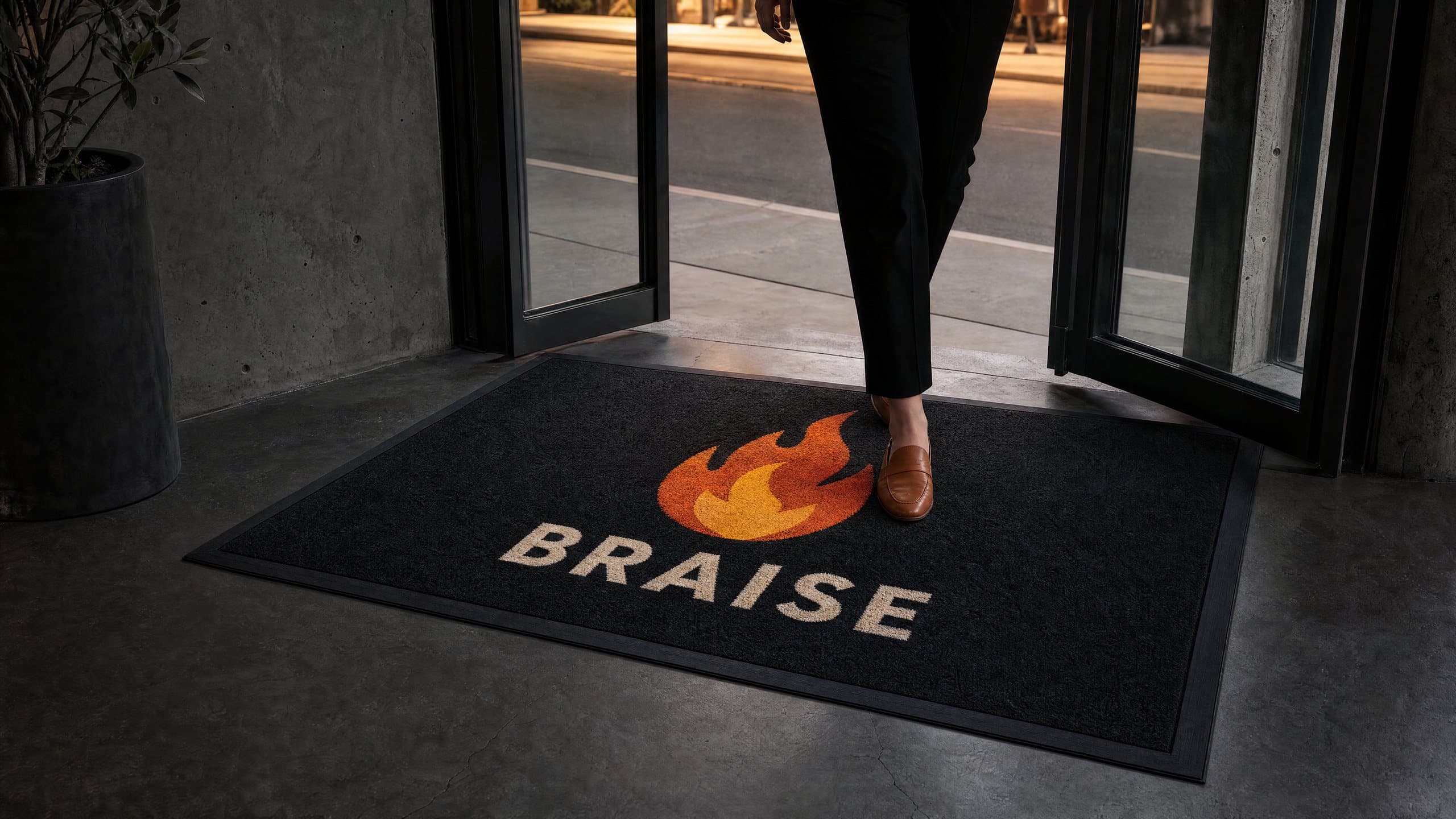 Tapis noir imprimé d’une flamme orange et du mot BRAISE à l’entrée vitrée d’un restaurant