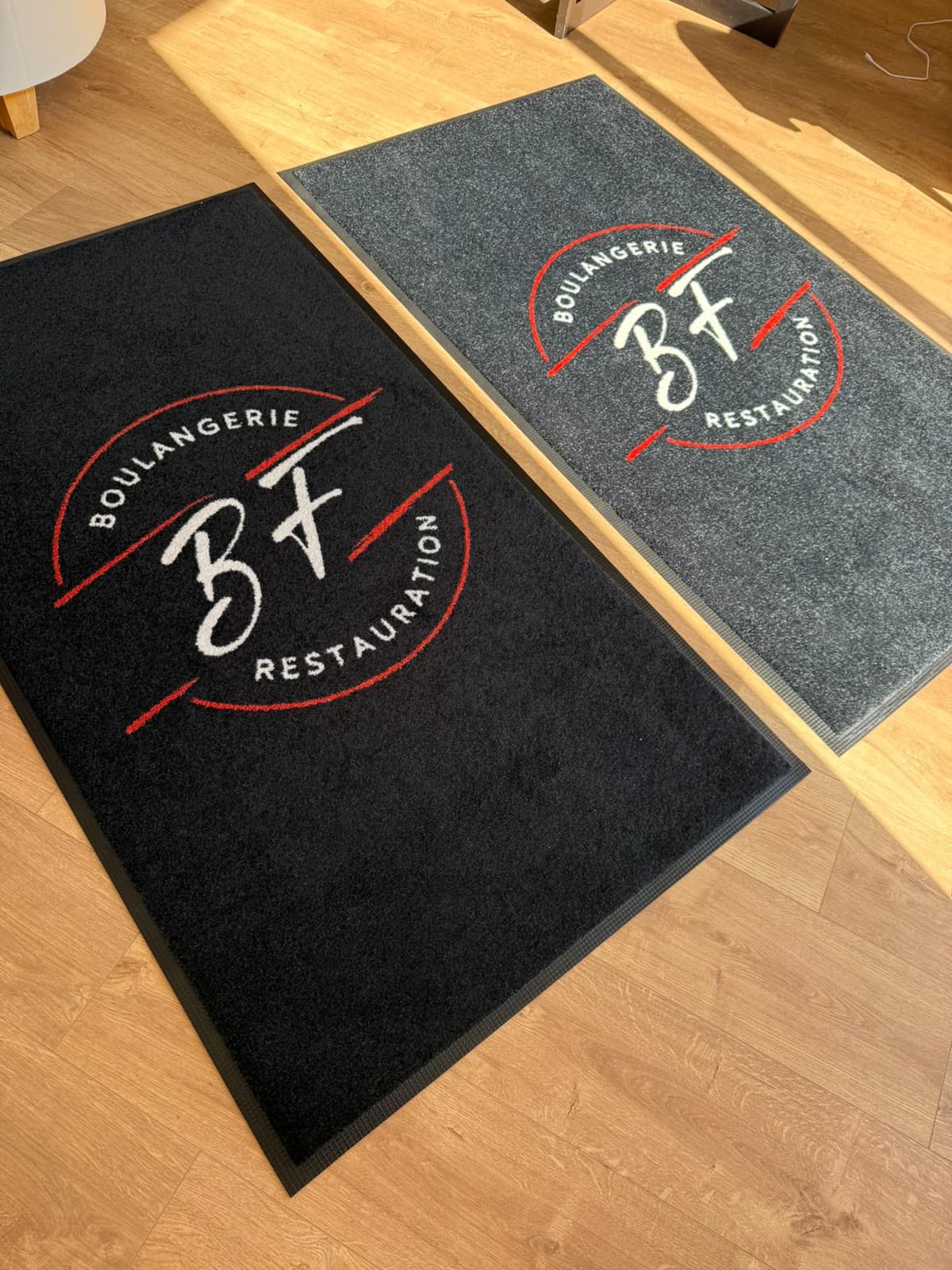 Deux tapis au logo BF Boulangerie Restauration, l’un noir, l’autre anthracite, posés côte à côte sur un parquet au soleil